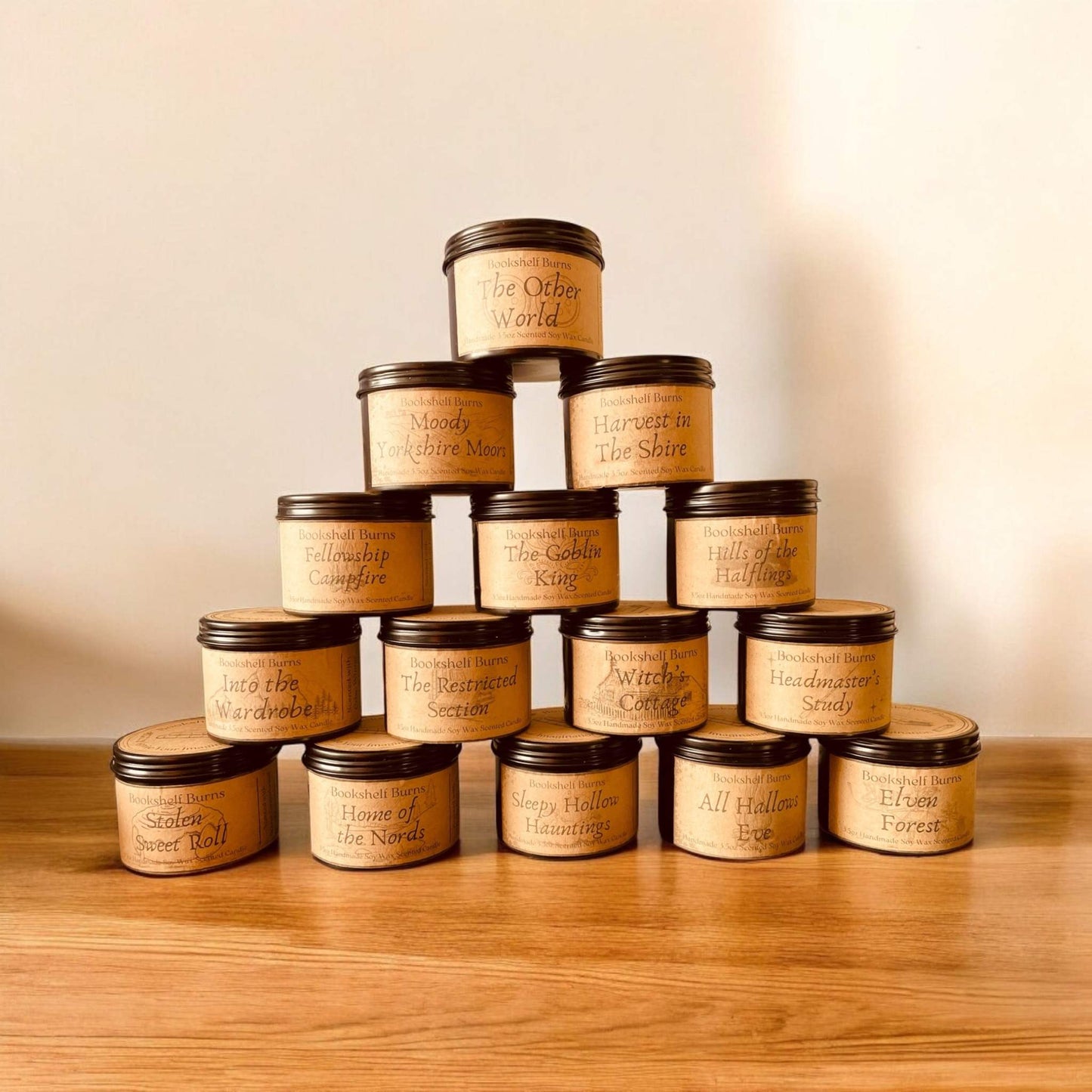 Hand poured scented candles stacked pyramid-style on a wooden surface, each labeled with literary-inspired names.