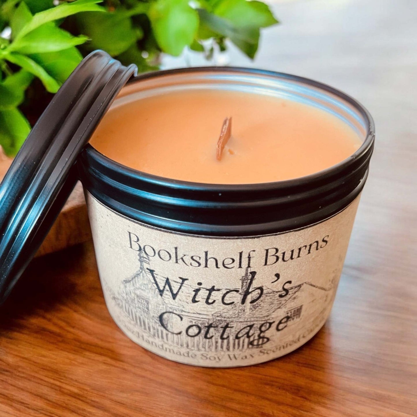 Hand-poured scented candle in a tin labeled "Witch's Cottage," set on a wooden surface with green leaves in the background.
